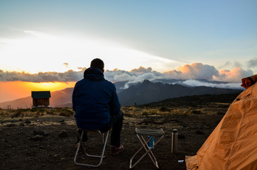 Hiker sitting on a folding chair at Shira Cave Camp, Mount Kilimanjaro, Tanzania, in the evening light next to a tent and an outhouse. Shira peak, westernmost peak of Kilimanjaro, in the background.