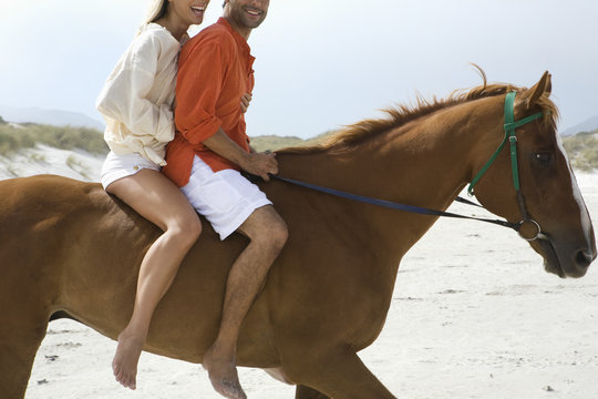 Side view of a cropped couple riding horse on beach