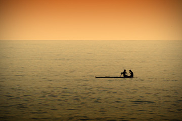 A couple resting while paddleboarding in a calm sea at sunset time. Dark orange atmosphere. Empty copy space for Editor's text.