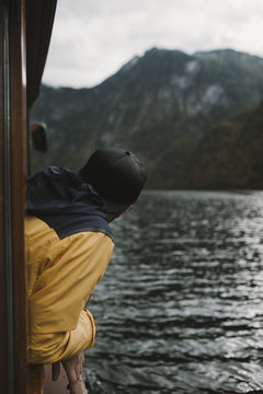Young adult male wearing yellow and blue jacket and cap looking out of a boat window on a mountain lake