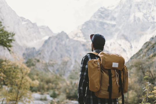 Young adult male wearing a flanel shirt cap and yellow backpack watching a mountain landscape with forest at bright daylight