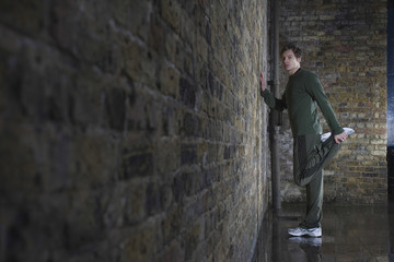 Fototapeta na wymiar Full length side view of a young man doing leg stretch against brick wall