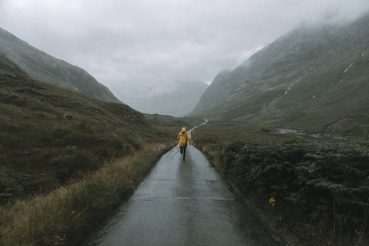 Man wearing a yellow jacket running down a road in a green field between mountains in the rain