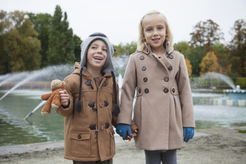 Portrait of happy brother and sister in trench coats holding hands at park