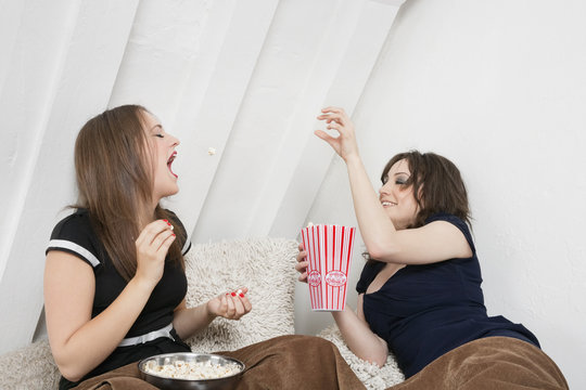 Playful Young Female Tossing Popcorn Into Friend's Mouth In Bed