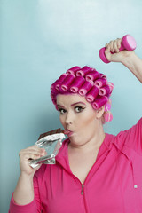 Portrait of young woman licking chocolate bar while light dumbbell over colored background