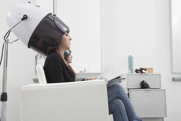 Side view of young woman under hooded dryer machine in hair salon