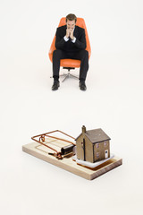 Model home on mouse trap with worried businessman sitting on chair representing increasing real...