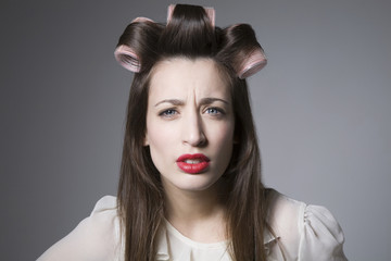 Portrait of a scowling young female with hair rollers and red lipstick isolated over grey background