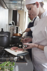 Female chef seasoning raw meat with colleague working in background at kitchen