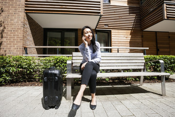 Full length of businesswoman answering cell phone while sitting by luggage on bench against building