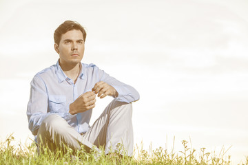 Thoughtful young man looking away while sitting on grass against clear sky