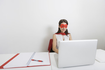 Young businesswoman dressed as superhero using laptop at desk in office