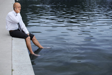 Fototapeta premium Businessman sitting on wall by lake, using mobile phone