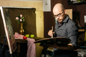 Artist draws a still life, holding a brush and palette. Apples and vase with flowers on background. In workroom