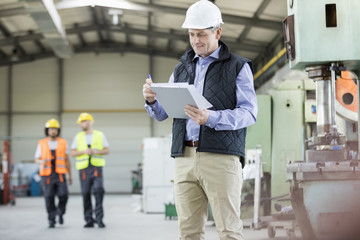 Mature male inspector writing on clipboard while workers in background at factory