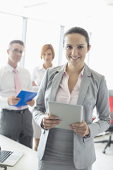 Portrait of happy businesswoman holding digital tablet with colleagues in background at office