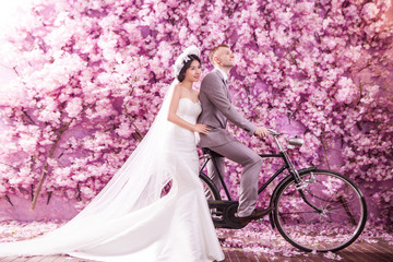 Fototapeta premium Thoughtful bride standing with bridegroom riding bicycle against pink flowers covering wall