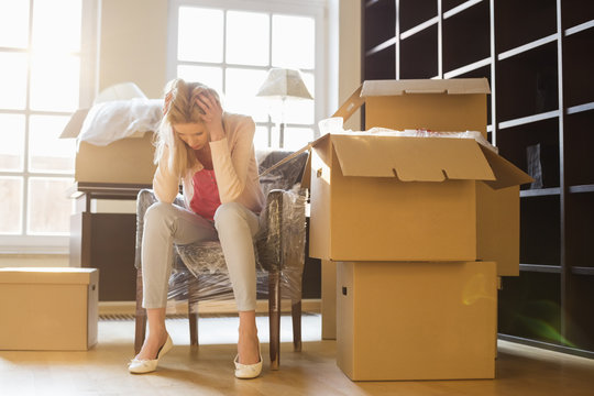 Full-length Of Frustrated Woman Sitting By Cardboard Boxes In New House