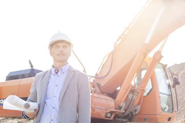 Thoughtful engineer looking away while holding blueprints by bulldozer at construction site