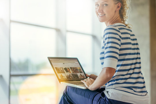 Rear view. Young smiling woman in striped T-shirt sitting and uses laptop. Girl browsing internet, chatting, blogging, checking email. Online shopping, learning. Window in soft focus on background.
