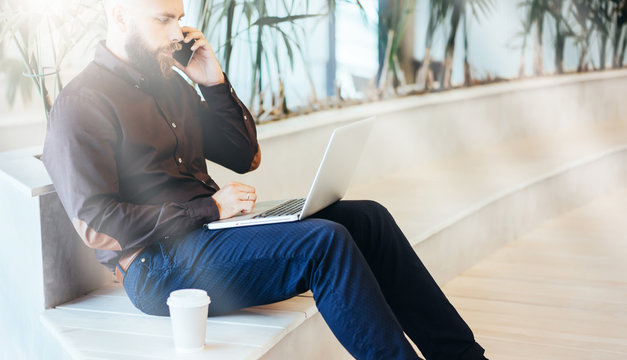 Side view. Young bearded businessman in brown shirt sitting and using laptop while talking on cell phone. Nearby stands cup of coffee. Man uses digital gadget. Guy chatting, blogging, checking email.