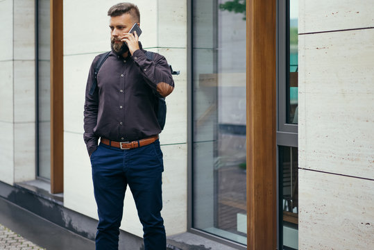 Young bearded businessman in a brown shirt with a backpack standing on a city street and talking on the cell phone. In the background is a modern building. Man using digital gadget.