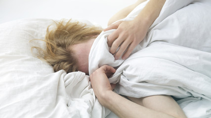 red-haired young man lying in bed covering up his face