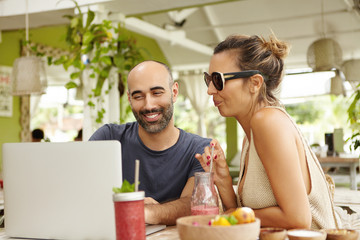 Obraz na płótnie Canvas Two people watching video online or viewing pictures on Internet, using wi-fi on laptop computer during lunch. Happy bearded man and stylish woman in shades relaxing at cafe with notebook. Film effect