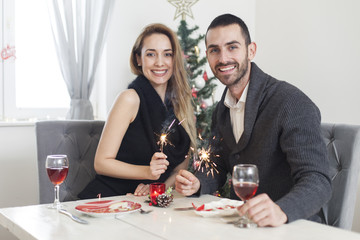 Young beautiful happy couple having a Christmas dinner holding fireworks sticks. New Years dinner