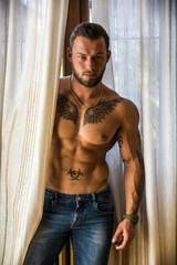 Portrait of sexy shirtless muscular man next to window curtains during the day, wearing only jeans