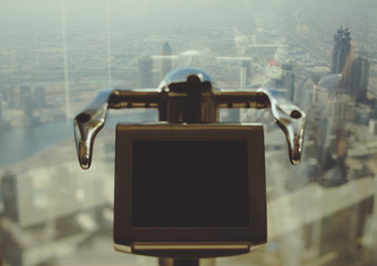 Fototapeta premium Tilt shift shooting of electronic telescope in front of window of Dubai skyscraper with top view of cityscape on summer day