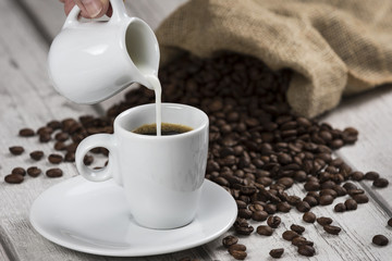 Coffee and Milk / Composition with cup of coffee and sack of coffee beans with hand pouring milk on a white wooden table