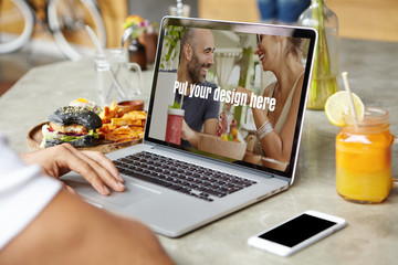 Young photographer working on open laptop, retouching photos sitting at table with food, orange juice and cell phone with blank screen for your design. Student doing home assignment on notebook