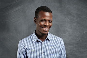Happy young dark-skinned man with small beard dressed in checkered shirt standing against blank chalkboard, looking at camera with cheerful smile. Human face expressions, emotions and feelings