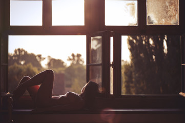 Beautiful naked woman sitting on a window at sunset