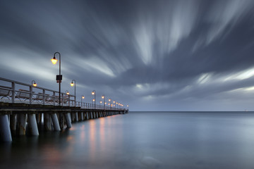 Calm Baltic Sea with illuminated pier and impressive stormy clouds on the sky. Early morning in Gdynia Orlowo. Poland. Europe. - obrazy, fototapety, plakaty