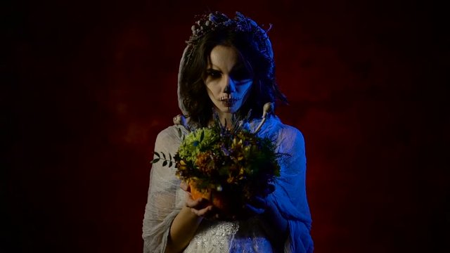 Close up of a ghost of young bride is standing with bunch of flowers in her hands. Mystical woman with art skull make-up on her face is staring at the camera and at the bunch.