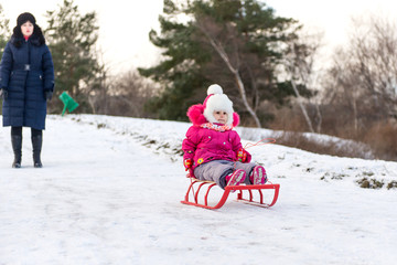 The child, a little girl riding on a sled with snow slides. Winter fun for children. A child with his mother and a winter walk.