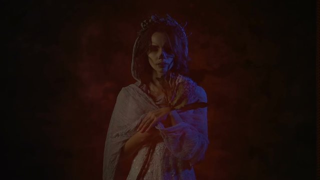 Ghost of a dead bride in white dress and wedding veil is appearing in the picture. Image of a young woman with skull mask on her face. Young creepy woman is standing with red background behind her.