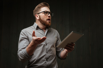 Surprised bearded man in eyeglasses holding tablet computer and gesturing
