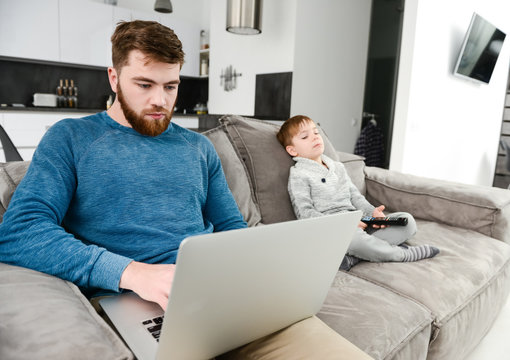 Serious bearded father using laptop while his son watching TV.