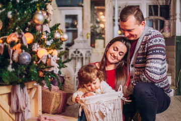 Fototapeta na wymiar Modern Christmas Family Portrait sitting and smiling In Home Holiday Living Room, Mother, father and baby girl .Present Gift Box, House Decorating By lights Xmas Tree Candles Garland. Box with gifts.