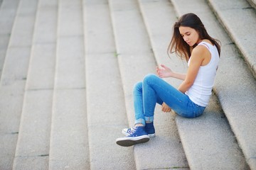 Fototapeta premium Young long-haired brunette girl in white tank top and blue jeans sitting pensively on the steps of stone stairs.