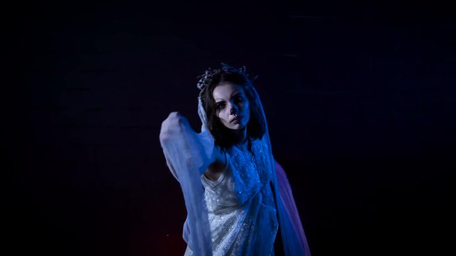 Ghost of young bride in white dress and veil. Moving scarry girl in with halloween make-up is standing against black background. Mysterious womanin horror behind .