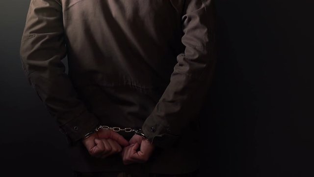 Arrested criminal cuffed man leaning on the wall in dark prison room, close up