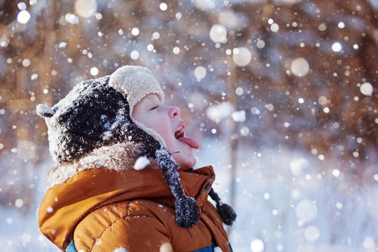Portrait of a cute little boy in warm clothes who catches snowflakes mouth in winter sunny day.