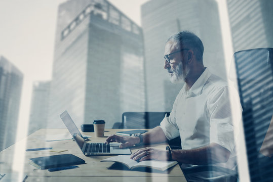 Double exposure concept.Adult bearded businessman working at wood table in modern place.Stylish middle age man using notebook.Skyscraper office building on the blurred background.Horizontal.