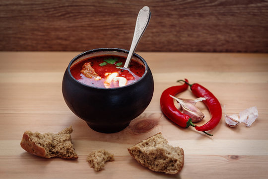 The soup is dark red in color. Soup in a cast iron pot. Board, which has chili pepper, garlic, bread, is a pot.