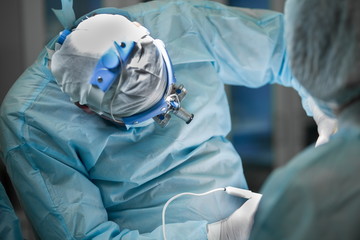 Surgeon and his assistant performing plastic surgery in hospital operating room. Surgeon in mask wearing loupes during medical procedure. Breast augmentation, enlargement, enhancement